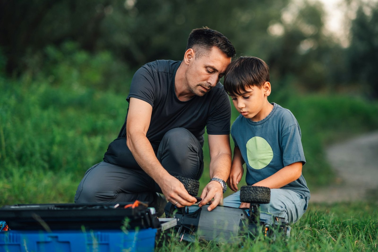 A caring father showing his son how to fix his electric toy car in nature.